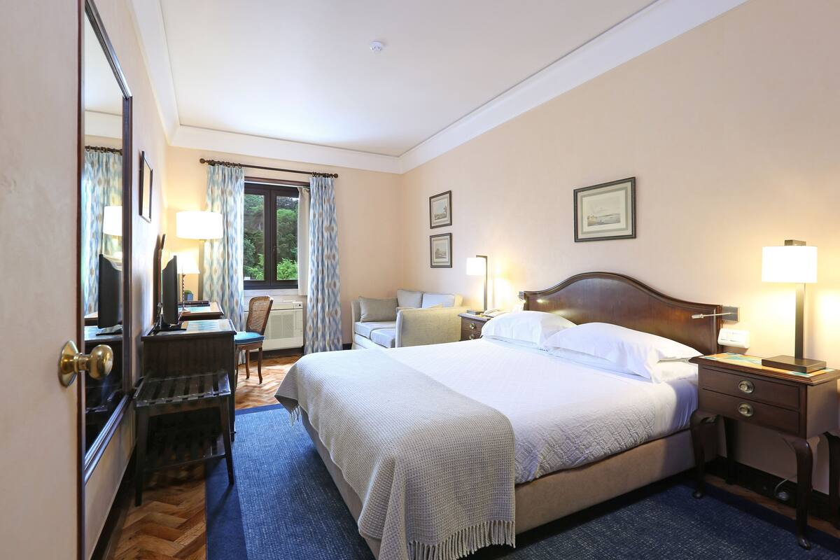 Habitación de hotel con una cama grande, mesitas de noche, un escritorio y una silla. Cuenta con ventanas con cortinas, una puerta y un espejo.