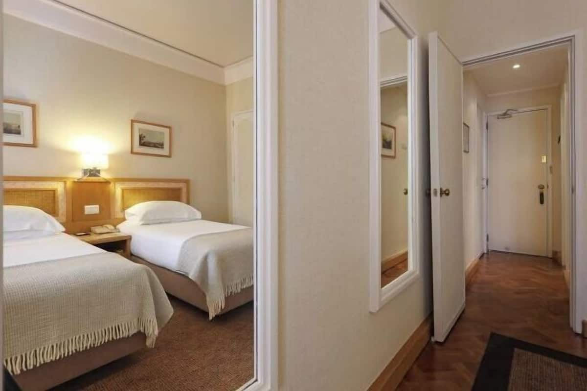 Habitación de hotel con dos camas, una mesita de noche, una lámpara y una puerta que da a otra habitación.