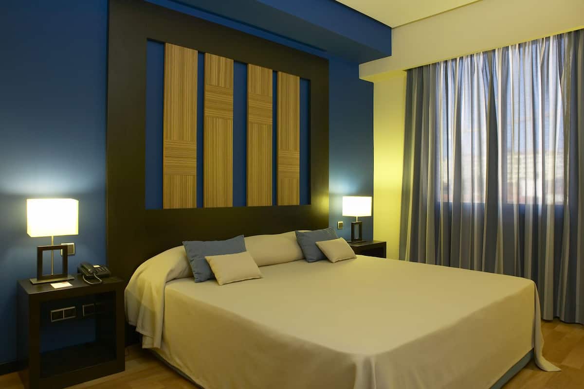 A hotel room with a large bed, two bedside tables with lamps, a wall-mounted artwork, and a window with curtains.