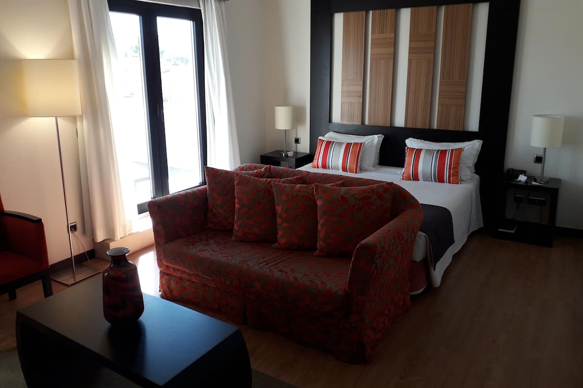A hotel room with a bed, a sofa, a nightstand, a lamp, and a window with curtains.