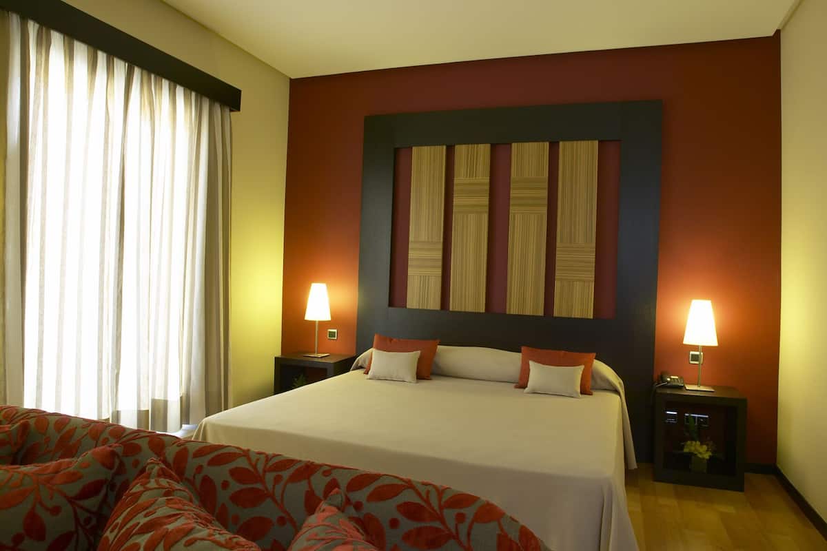 A hotel room with a large bed, two bedside tables with lamps, a window with curtains, and a floral-patterned sofa.
