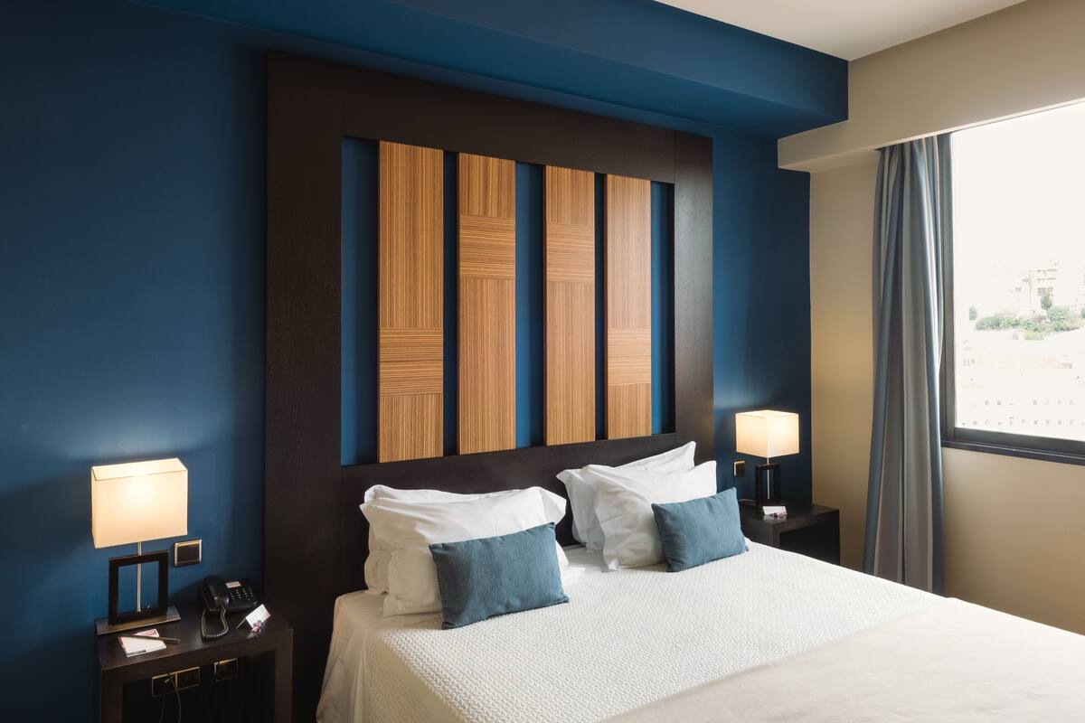 A hotel room with a large bed, two bedside tables with lamps, a wooden headboard, and a window with curtains.