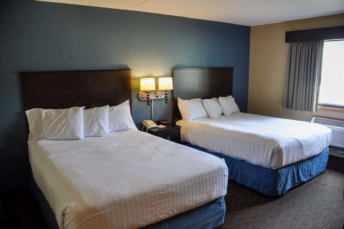 A hotel room with two beds, a nightstand with a lamp, and a window with curtains.