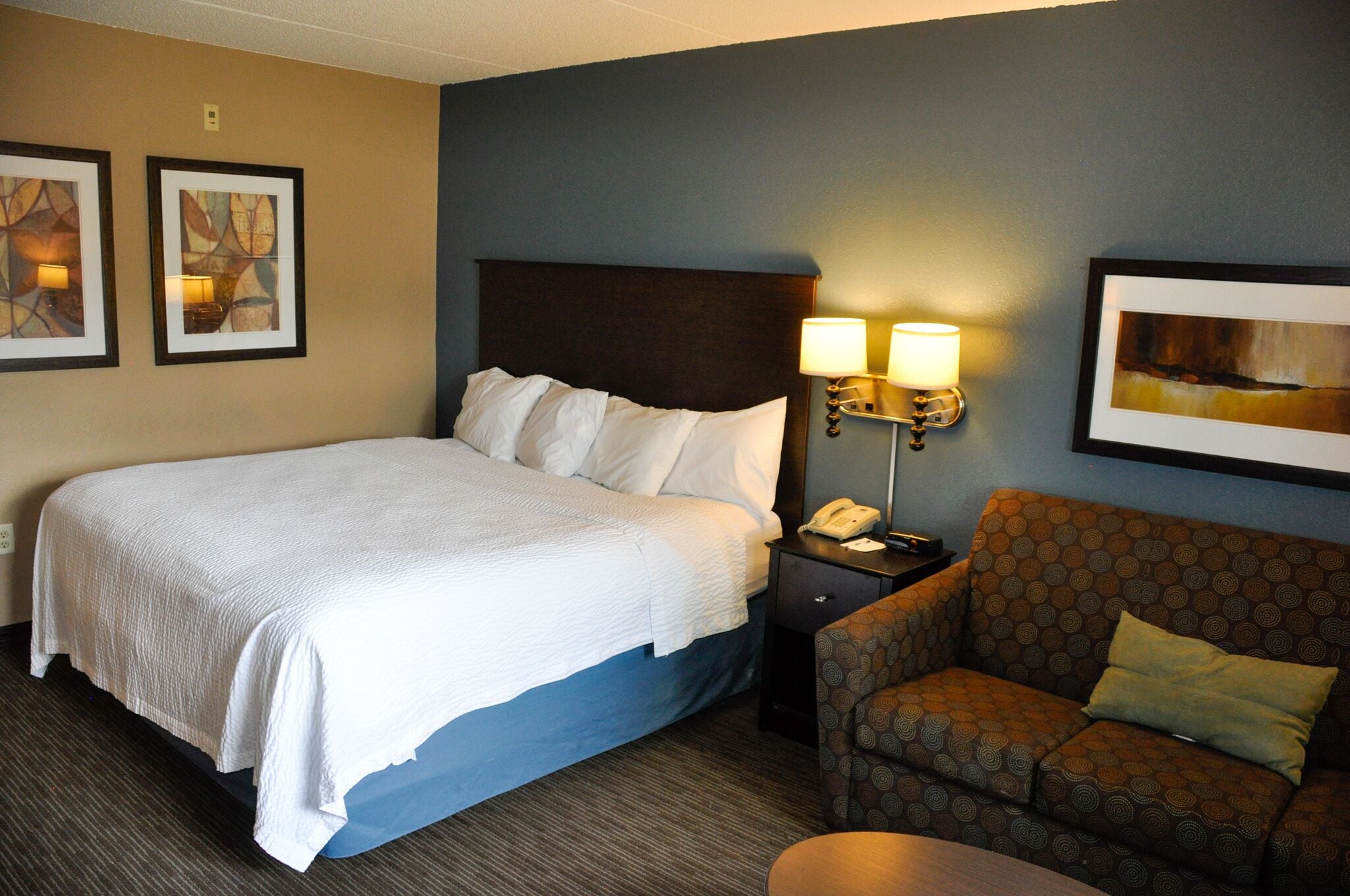 A hotel room with a large bed, a nightstand with a lamp, a sofa, and two framed artworks on the wall.