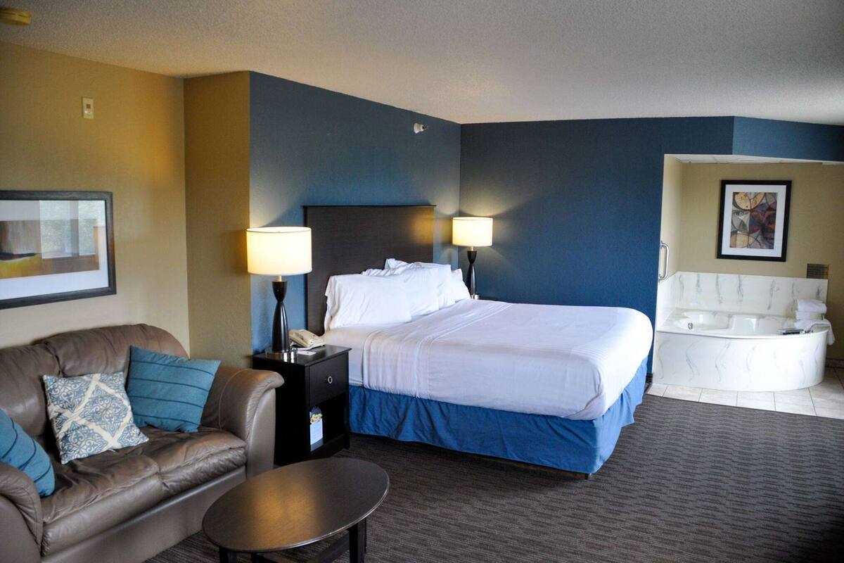 A hotel room with a bed, a sofa, a nightstand, a lamp, a table, and a bathtub.