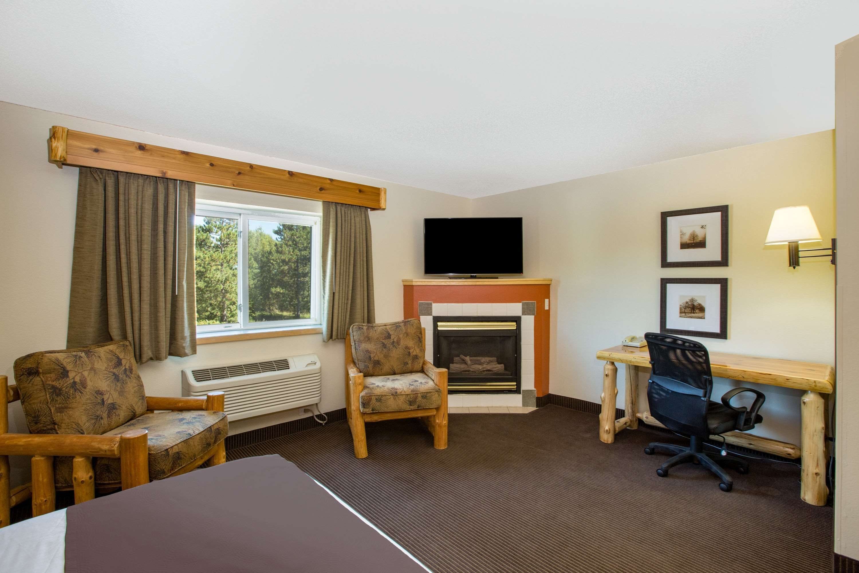 A hotel room with a bed, two armchairs, a desk with a computer, a fireplace, a TV, and a window with curtains.