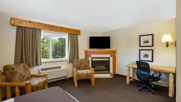 A hotel room with a bed, two armchairs, a desk with a computer, a fireplace, a TV, and a window with curtains.