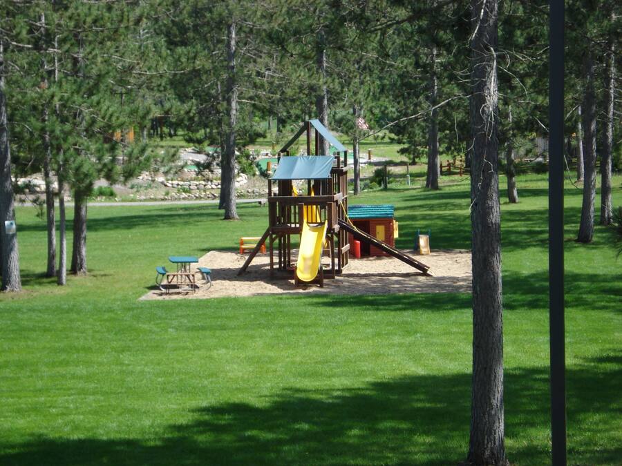 A playground with a slide, climbing structure, and sand area, surrounded by trees and grass.