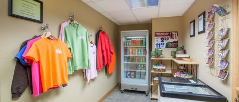 A clothing store with racks of colorful shirts, a refrigerated display case, and a wall with framed pictures and a sign.