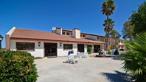Un bâtiment à deux étages, doté d’un toit recouvert de tuiles rouges, d’un patio avec des chaises en plastique blanches et d’un palmier en arrière-plan.