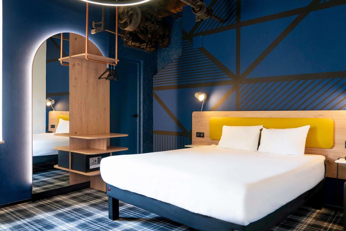 Ein modernes Hotelzimmer mit einem großen Bett, einem hölzernen Kopfteil, einem Spiegel und einer Wand mit geometrischem Muster.
