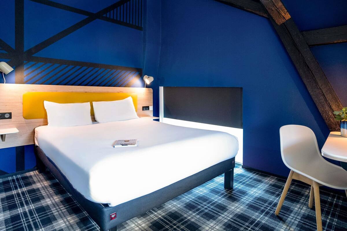 A modern hotel room with a bed, a desk, a chair, and a blue accent wall.