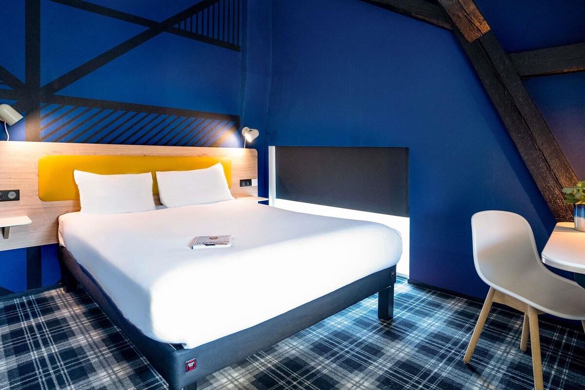 A modern hotel room with a bed, a desk, a chair, and a blue accent wall.