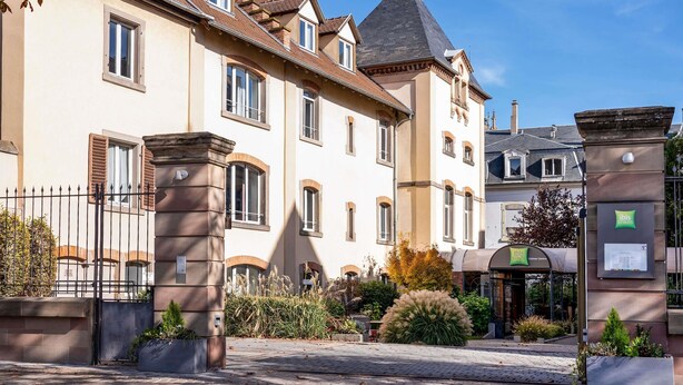 A historic building with a prominent tower, a gated entrance, and a nearby modern structure with a sign. - ibis Styles Colmar Centre (Colmar)