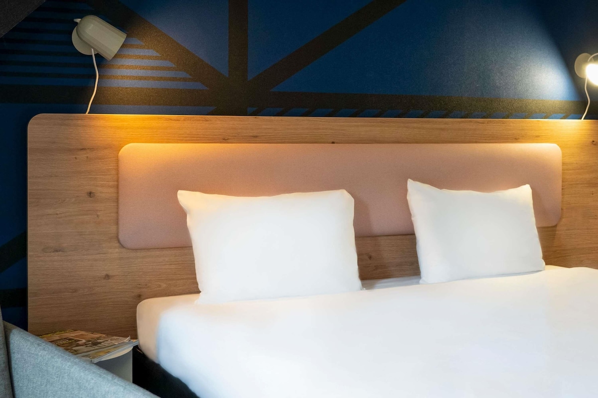 A neatly made bed with white bedding and two pillows, a wooden headboard, and a wall with a geometric pattern.