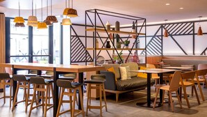 A modern cafe with wooden tables and chairs, a central seating area with a sofa, and a geometric wall design. - ibis Styles Colmar Centre (Colmar)
