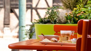 A table with a mug, bread, and a plate of food, a green planter with plants, and an orange chair. - ibis Styles Colmar Centre (Colmar)