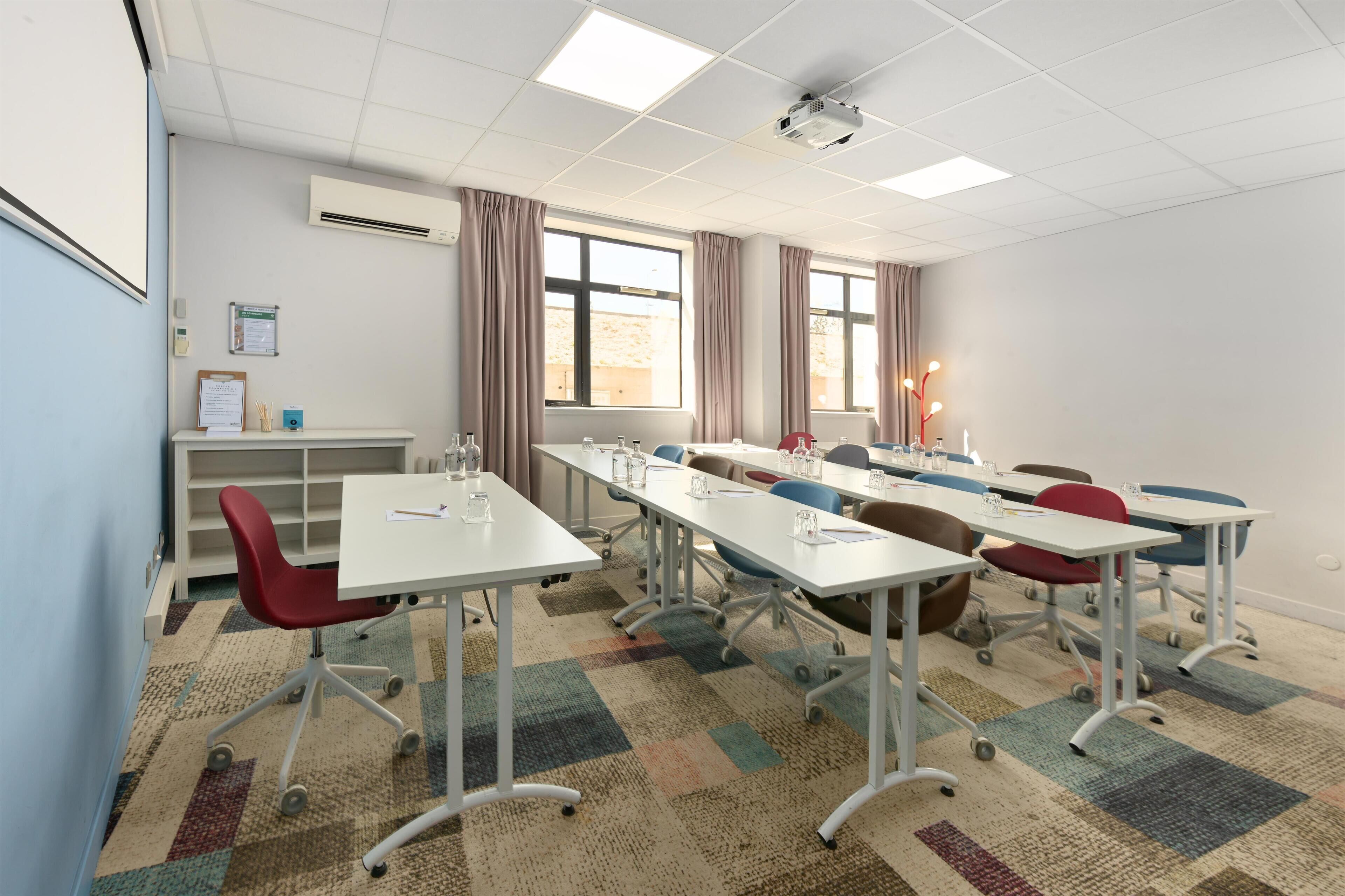 A conference room with white tables, red and blue chairs, a projector, and a carpeted floor.