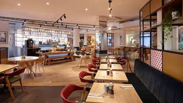L’intérieur d’un restaurant avec des tables en bois, des chaises rouges, un bar et un espace salon agrémenté d’un banc noir rembourré.