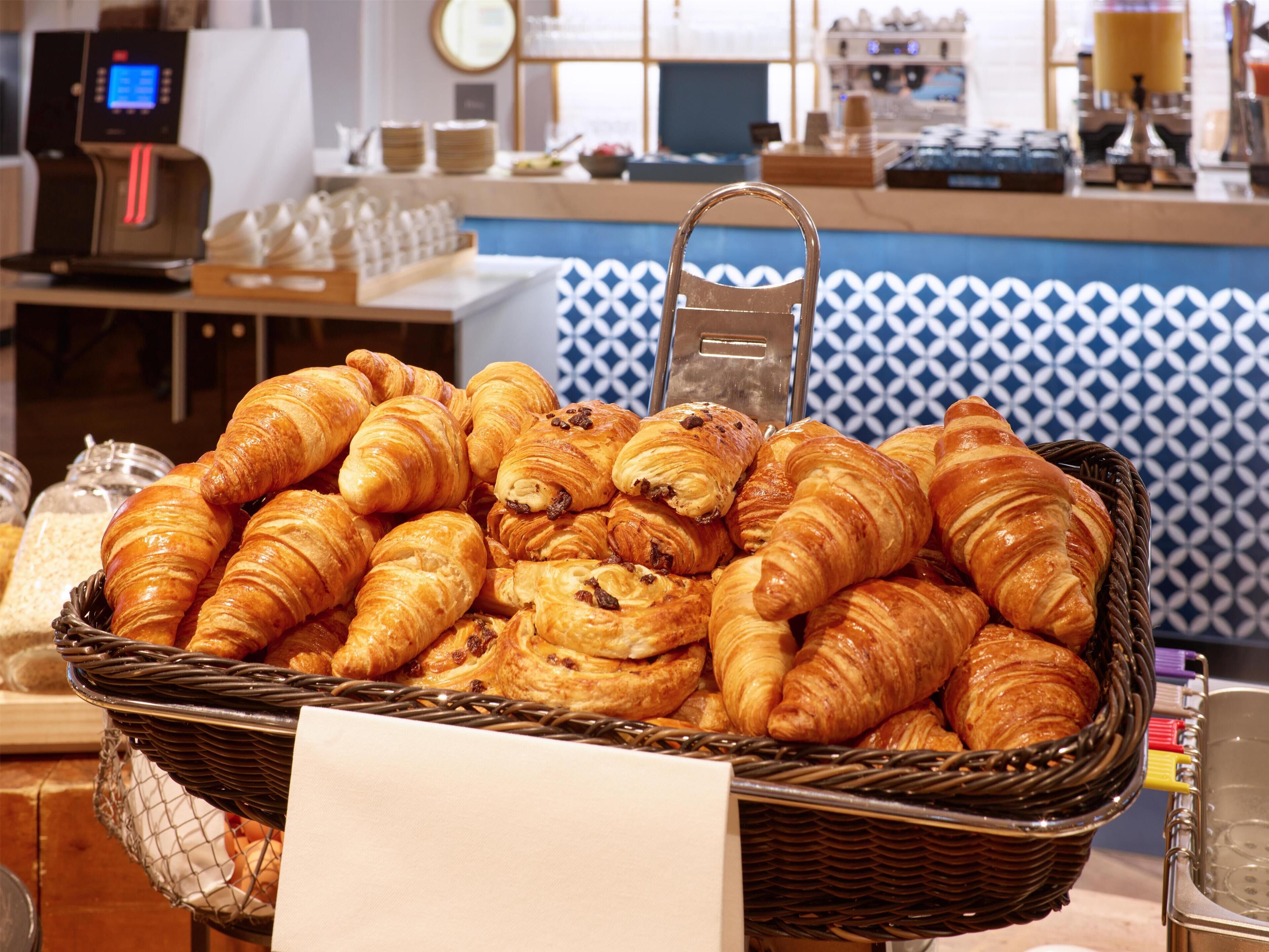 A basket of assorted pastries, including croissants and chocolate-filled pastries, displayed in a café setting with a blue and white patterned wall in the background.