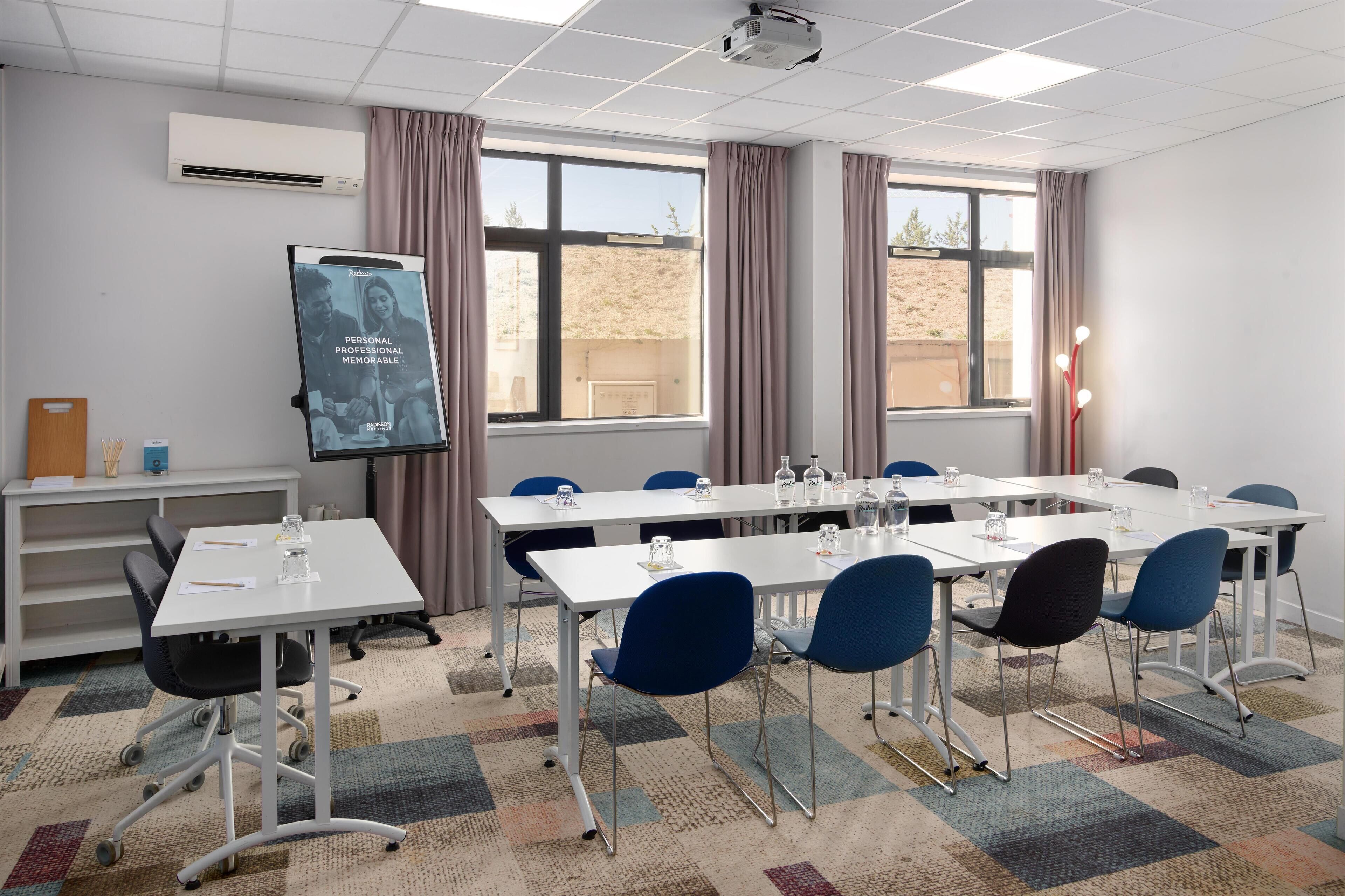 A conference room with a large table, chairs, a projector screen, and a window with curtains.