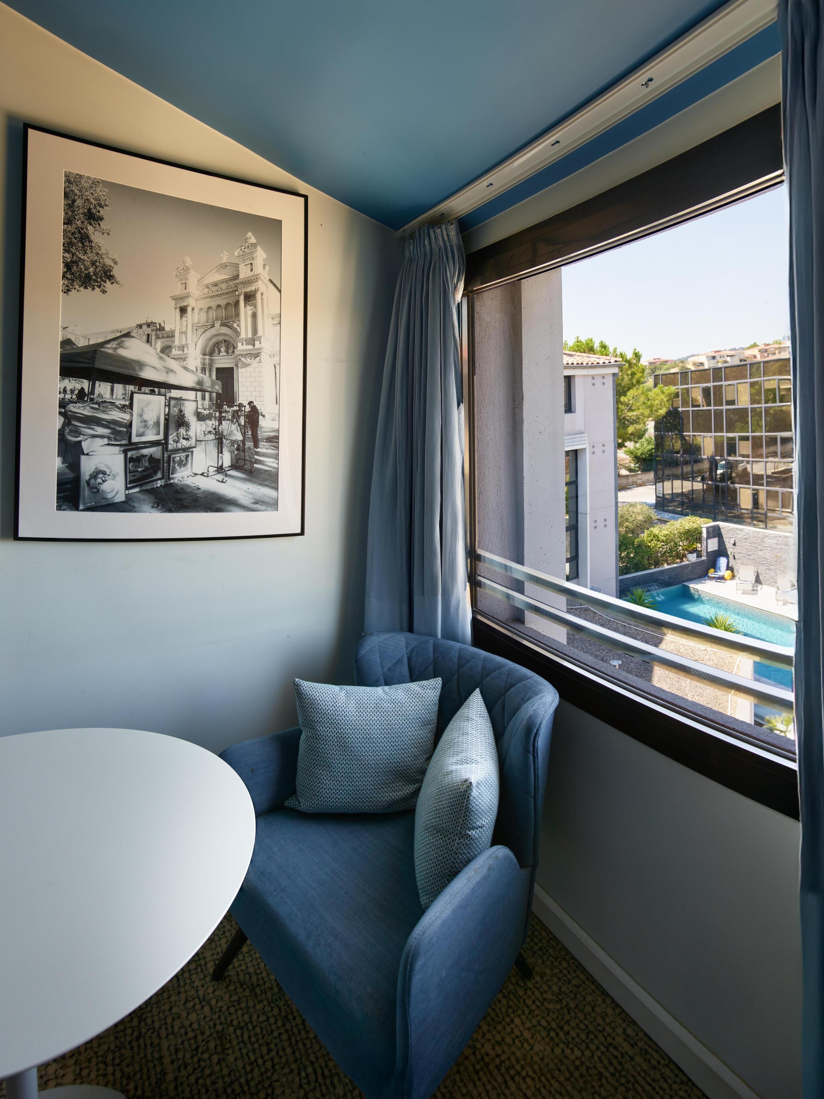 A room with a blue armchair, a white table, a framed black and white photo, and a view of a swimming pool through a window.