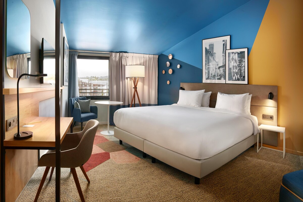 A modern hotel room with a large bed, a desk, a chair, a small table, and a view of the city.