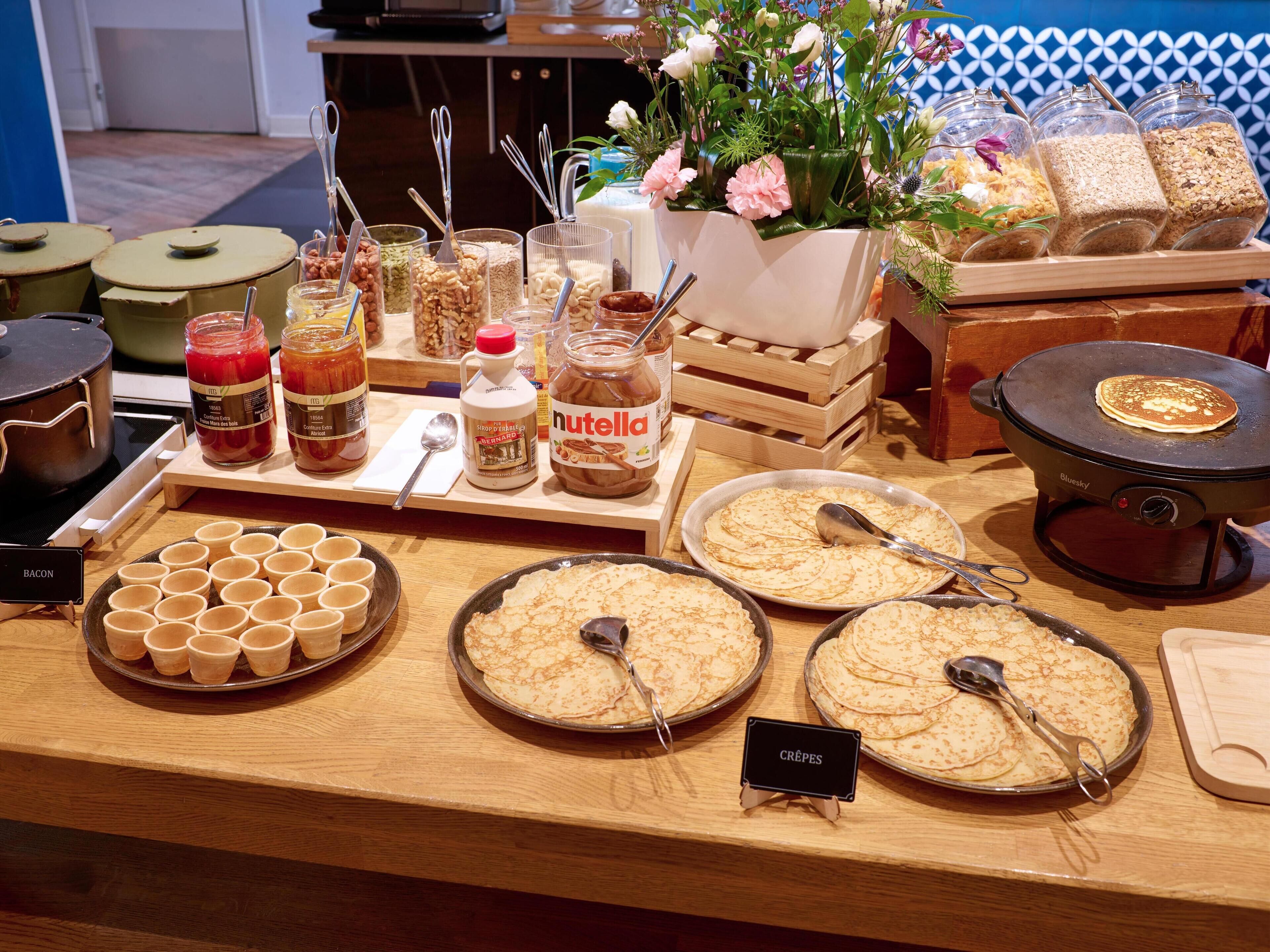 Una mesa de buffet con distintos alimentos, incluyendo frascos de conservas, un frasco de Nutella y varias porciones de crepes.