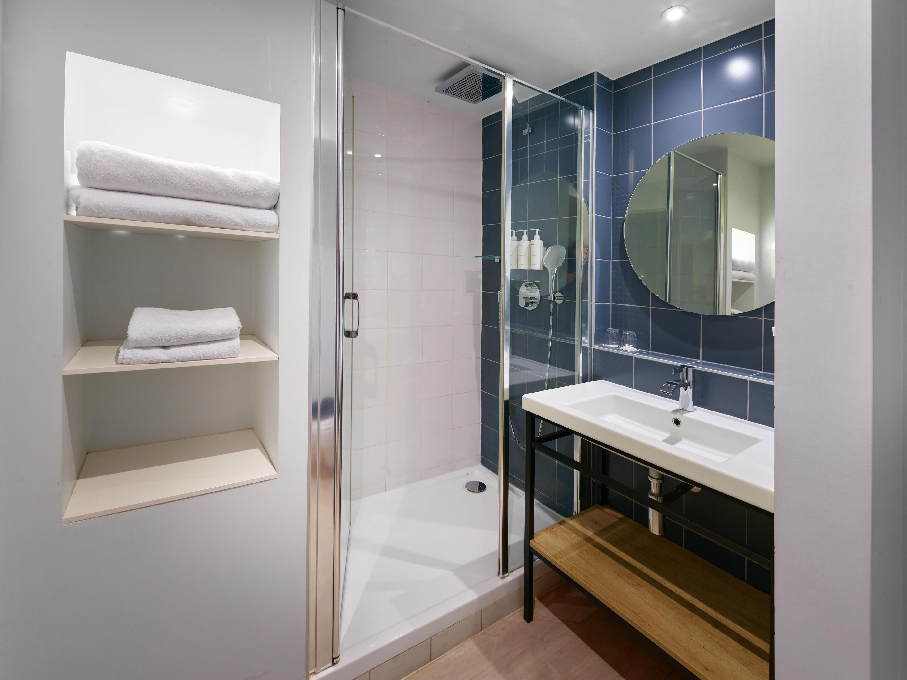 A modern bathroom with a glass-enclosed shower, white shelves with towels, a sink, and a mirror.