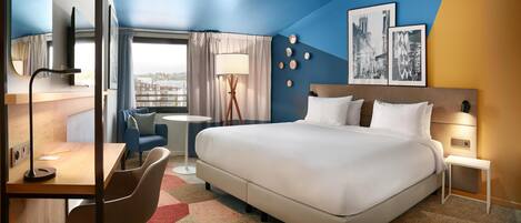 A modern hotel room with a large bed, a desk, a chair, a small table, and a view of the city.
