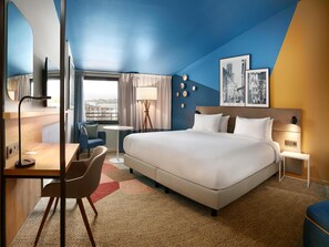 A modern hotel room with a large bed, a desk, a chair, a small table, and a view of the city. - Radisson Hotel Aix en Provence (Aix-en-Provence)