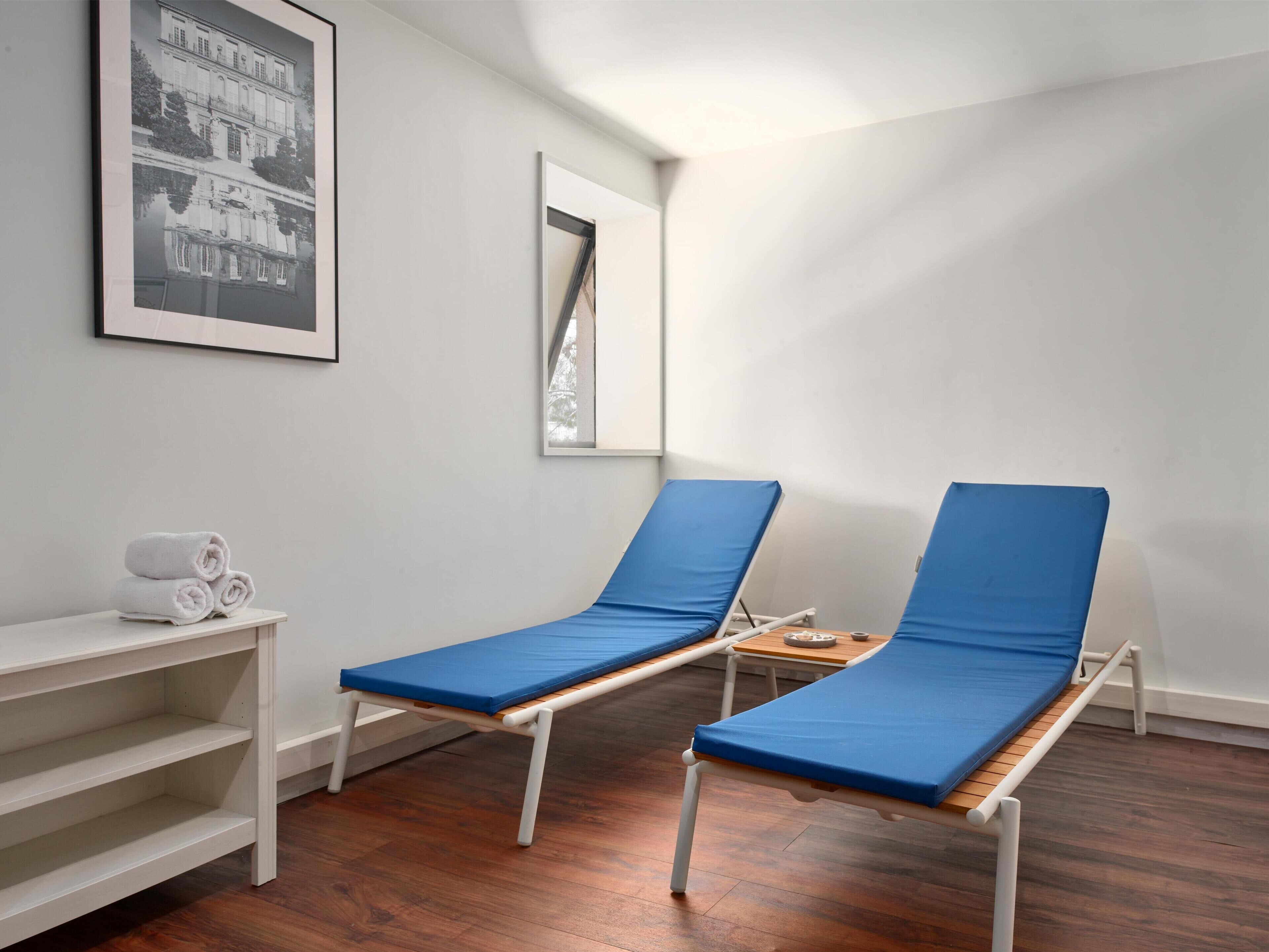 Ein Zimmer mit zwei blauen Polstersesseln, einem weißen Regal mit Handtüchern und einem gerahmten Bild an der Wand.