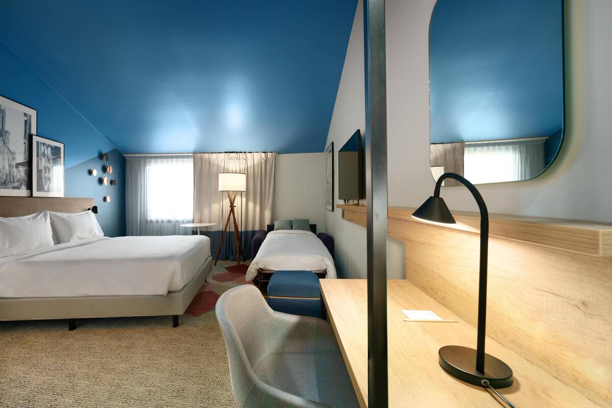 A hotel room with two beds, a desk, a chair, a lamp, a TV, and a large mirror.