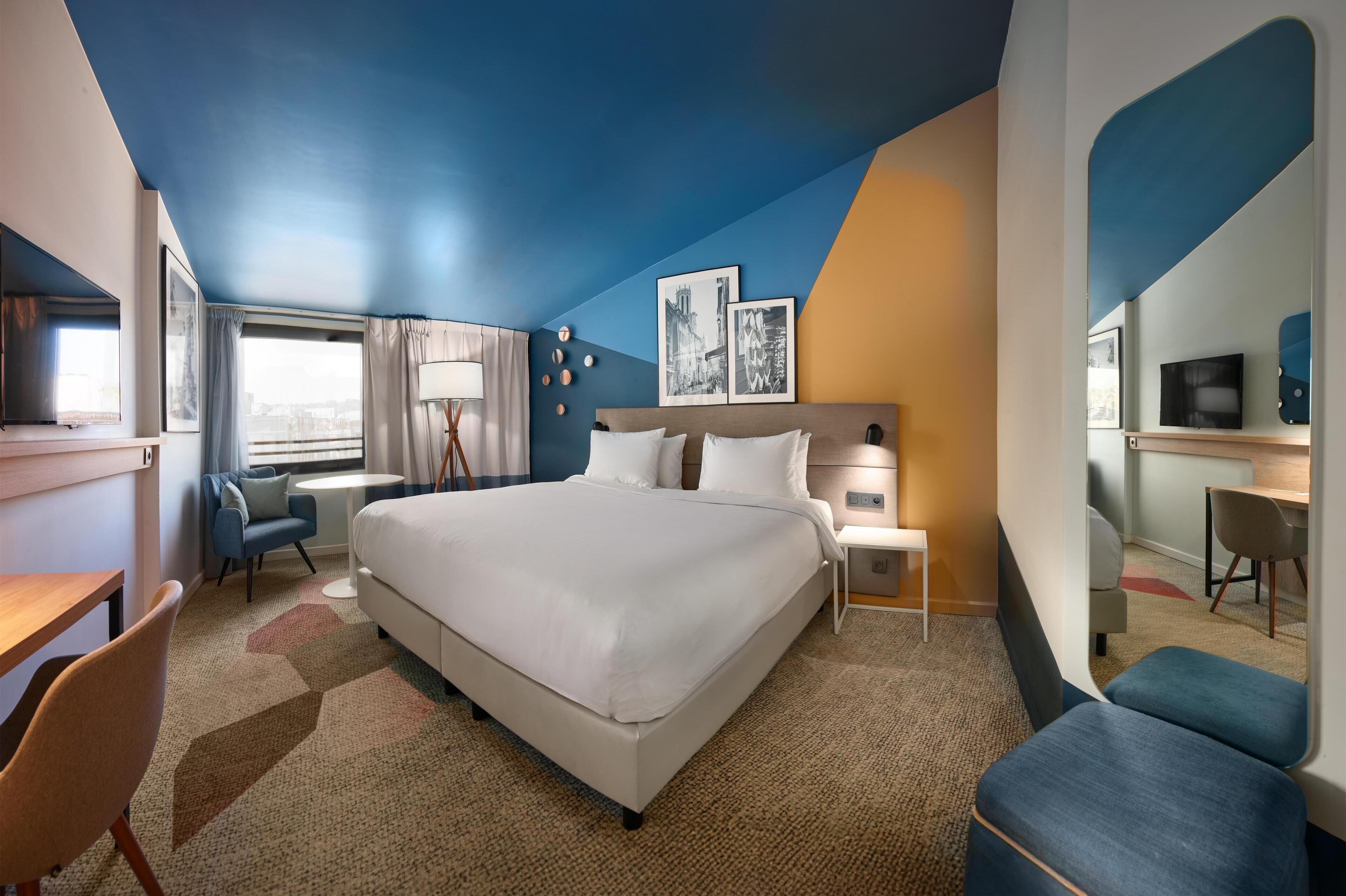 Een hotelkamer met een groot bed, een bureau, een stoel en een kleine tafel. Er is een raam met gordijnen, een lamp en een aan de muur bevestigde televisie.