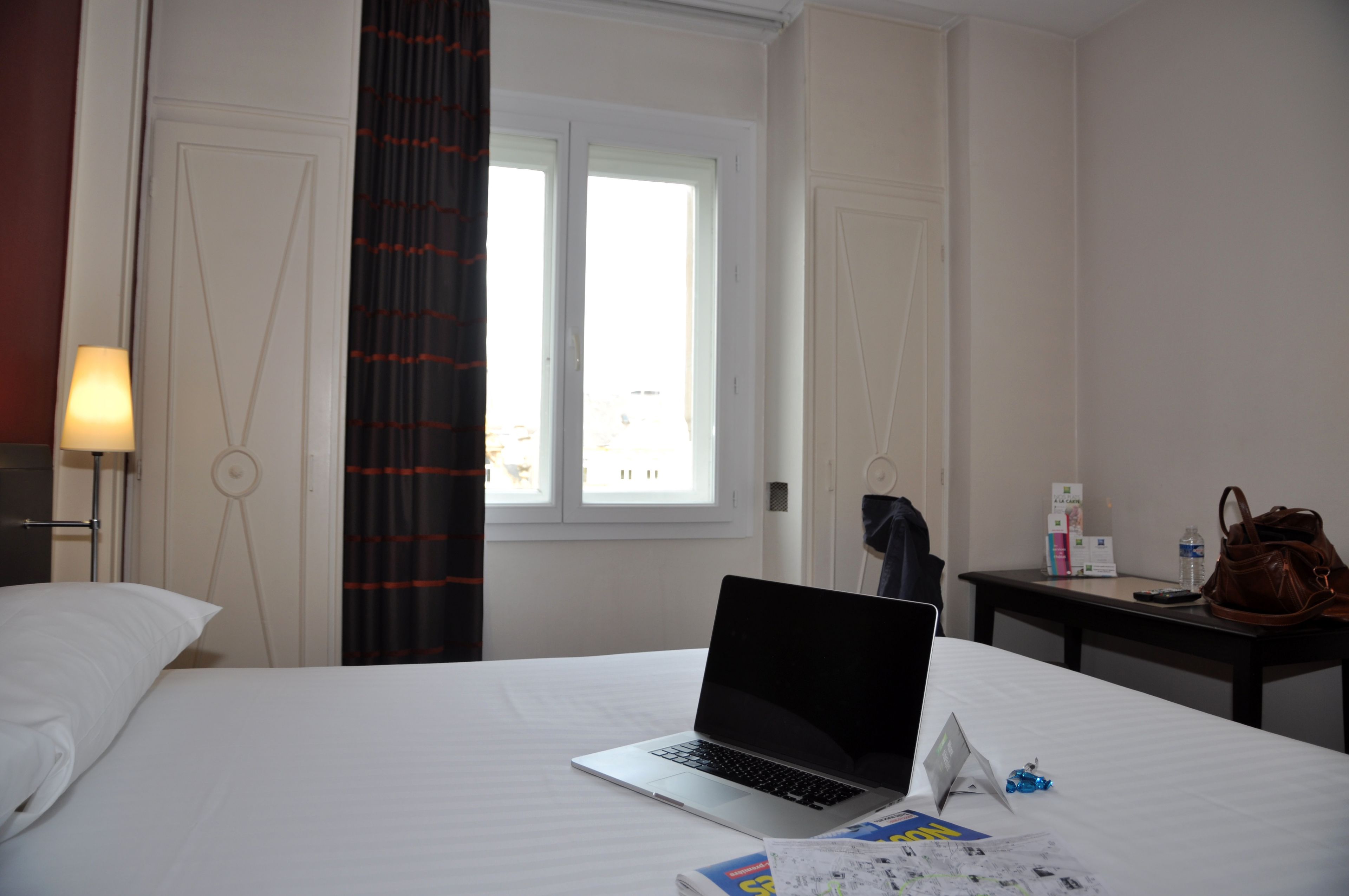 Una habitación de hotel con cama, laptop, mesita de noche con lámpara, ventana y puerta.