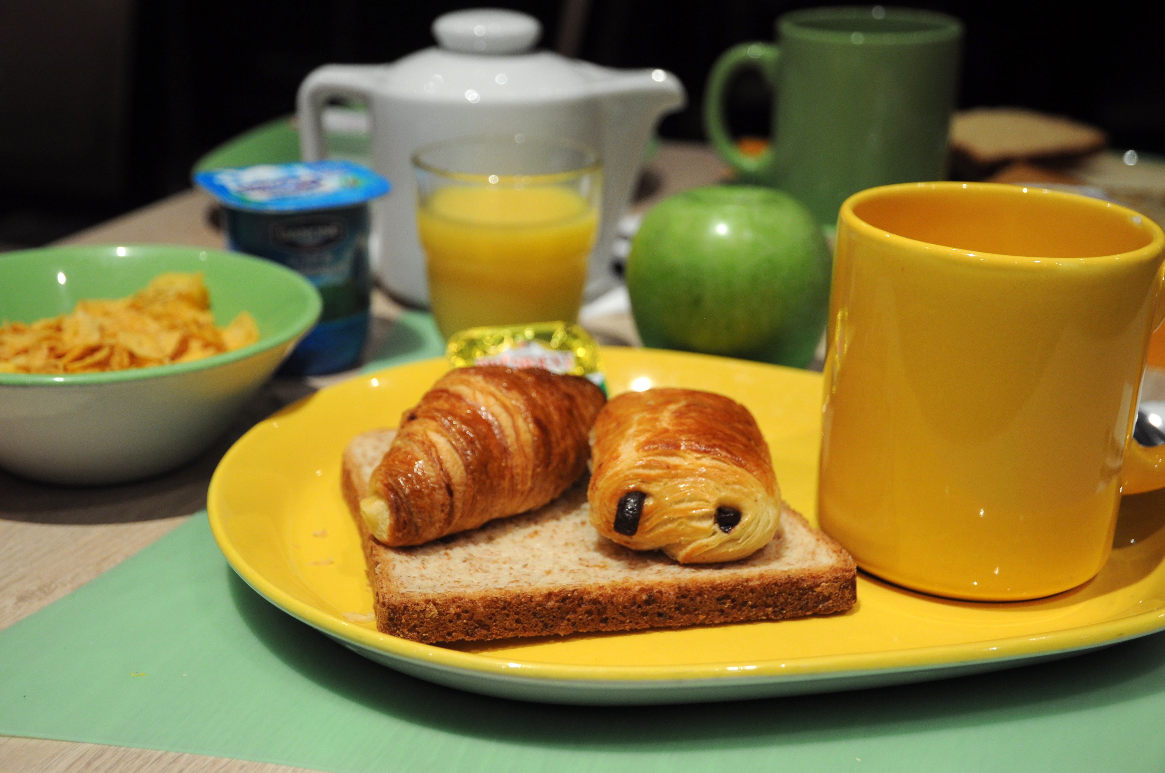 A breakfast setting with croissants, toast, a mug, and a bowl of cereal on a yellow plate.