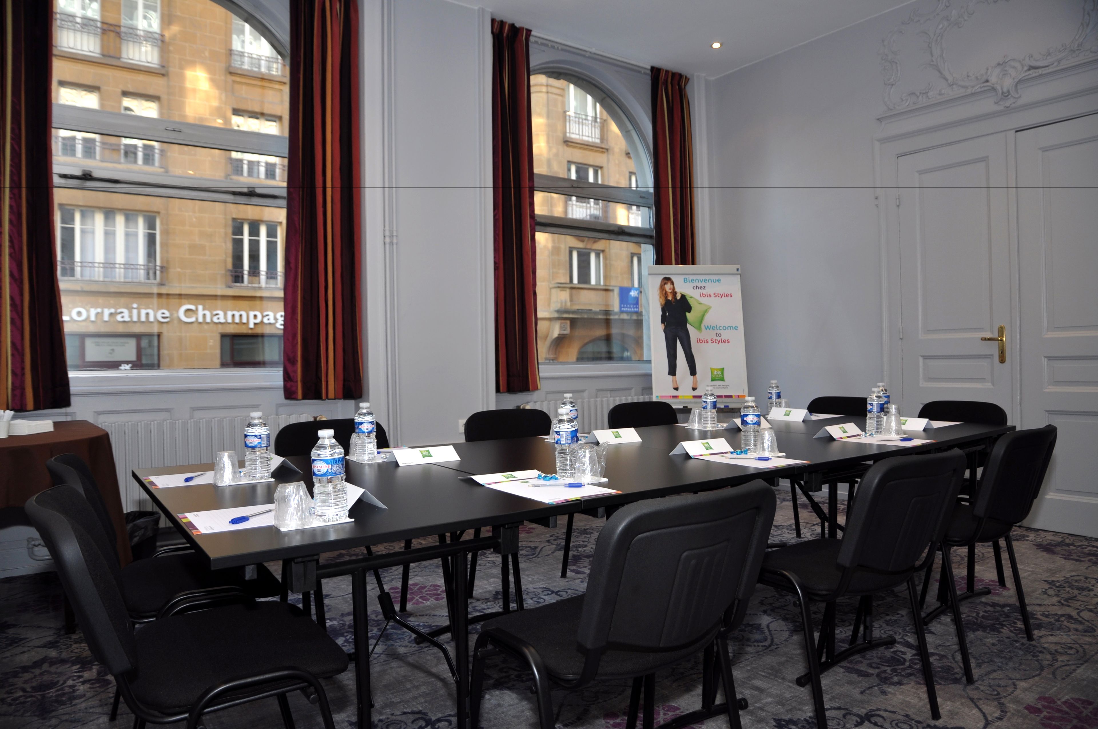 Sala de conferência com uma mesa comprida, cadeiras pretas, garrafas de água e tela de apresentação.