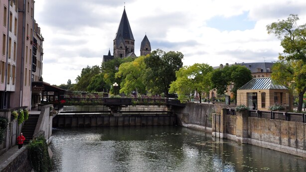 A river with a bridge, a church with a tall spire, and buildings along the riverbank. - ibis Styles Metz Centre Gare (Metz)