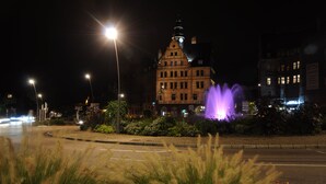A nighttime cityscape with a prominent building featuring a clock tower, illuminated by streetlights and a purple fountain. - ibis Styles Metz Centre Gare (Metz)