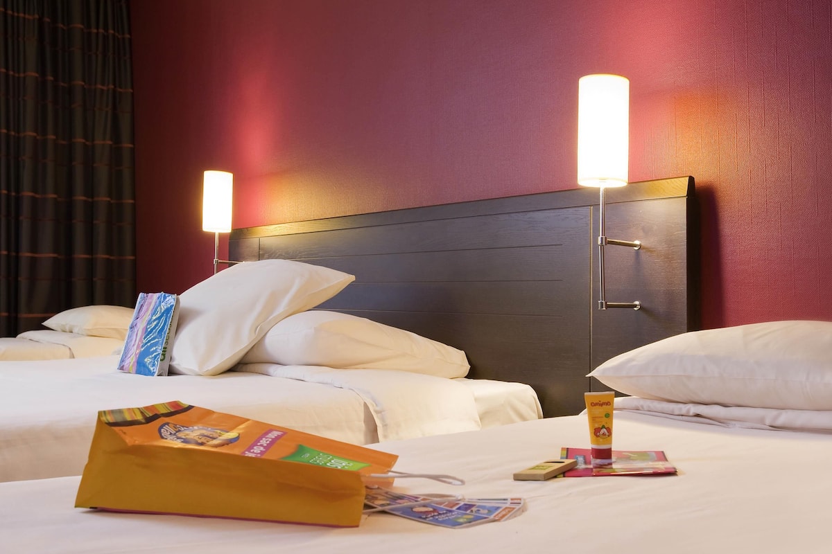 A hotel room with two beds, a wooden headboard, bedside lamps, and a bag of toiletries on the bed.