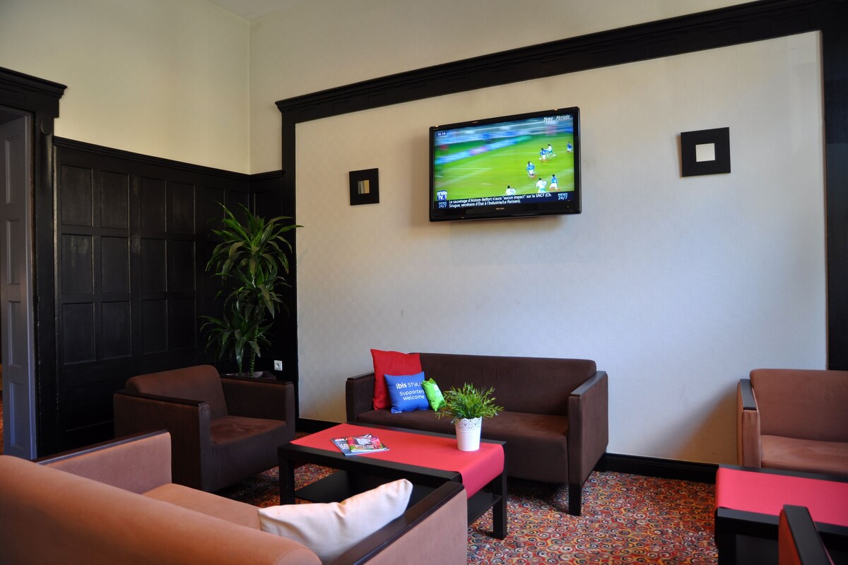 A modern lounge area with a flat-screen TV mounted on the wall, a brown sofa with a red cushion, a small table with a plant, and a colorful patterned carpet.