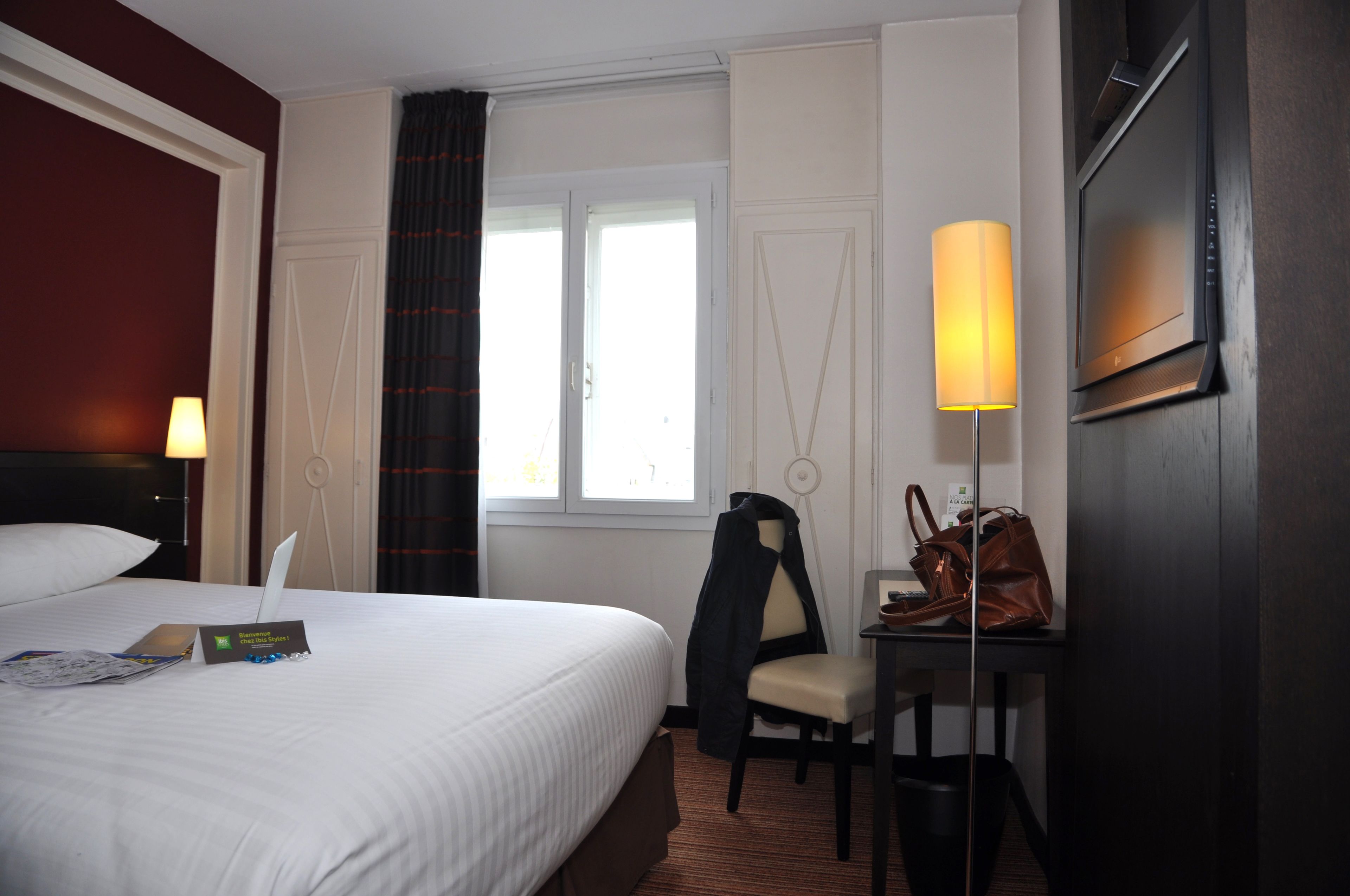 A hotel room with a bed, a desk with a lamp, a chair, a TV, and a window with curtains.