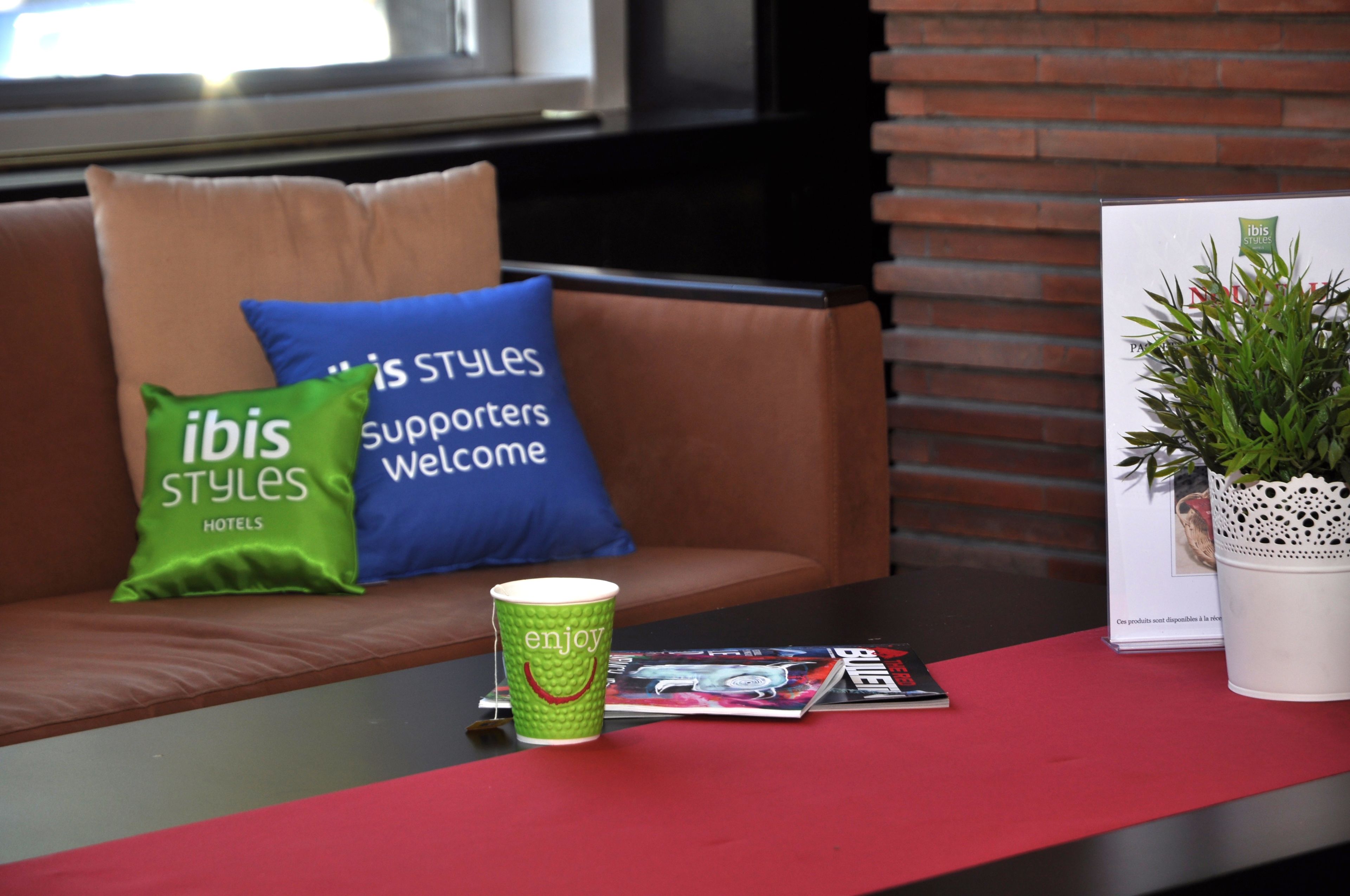 A seating area with a brown sofa, green and blue pillows, a green cup with 'enjoy' text, a magazine, and a white potted plant.