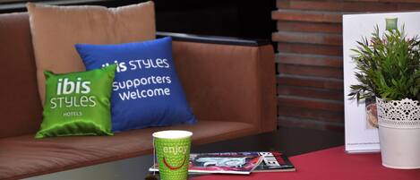 A seating area with a brown sofa, green and blue pillows, a green cup with 'enjoy' text, a magazine, and a white potted plant.