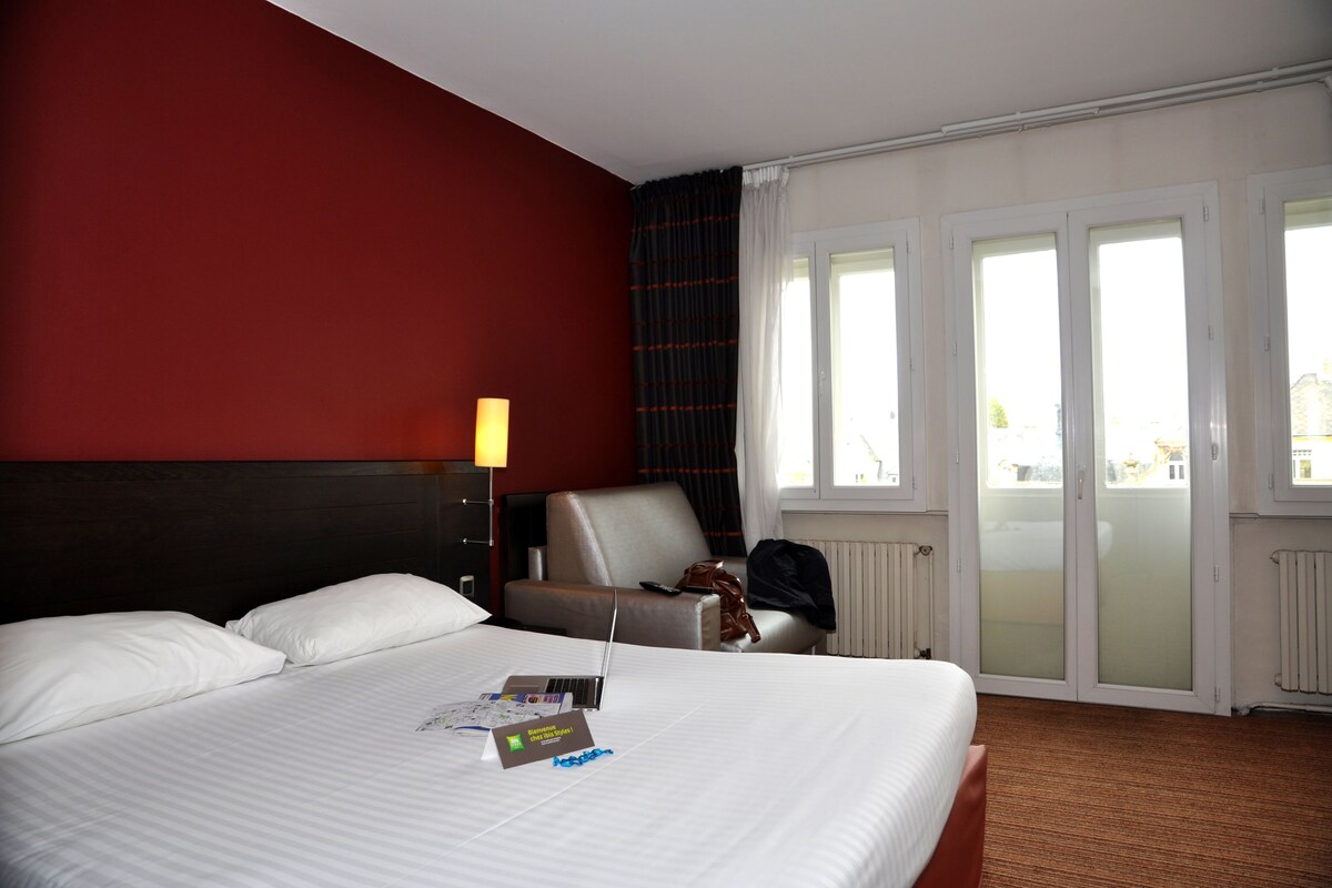 A hotel room with a large bed, a chair, a desk, and a view of the outside through a sliding door.