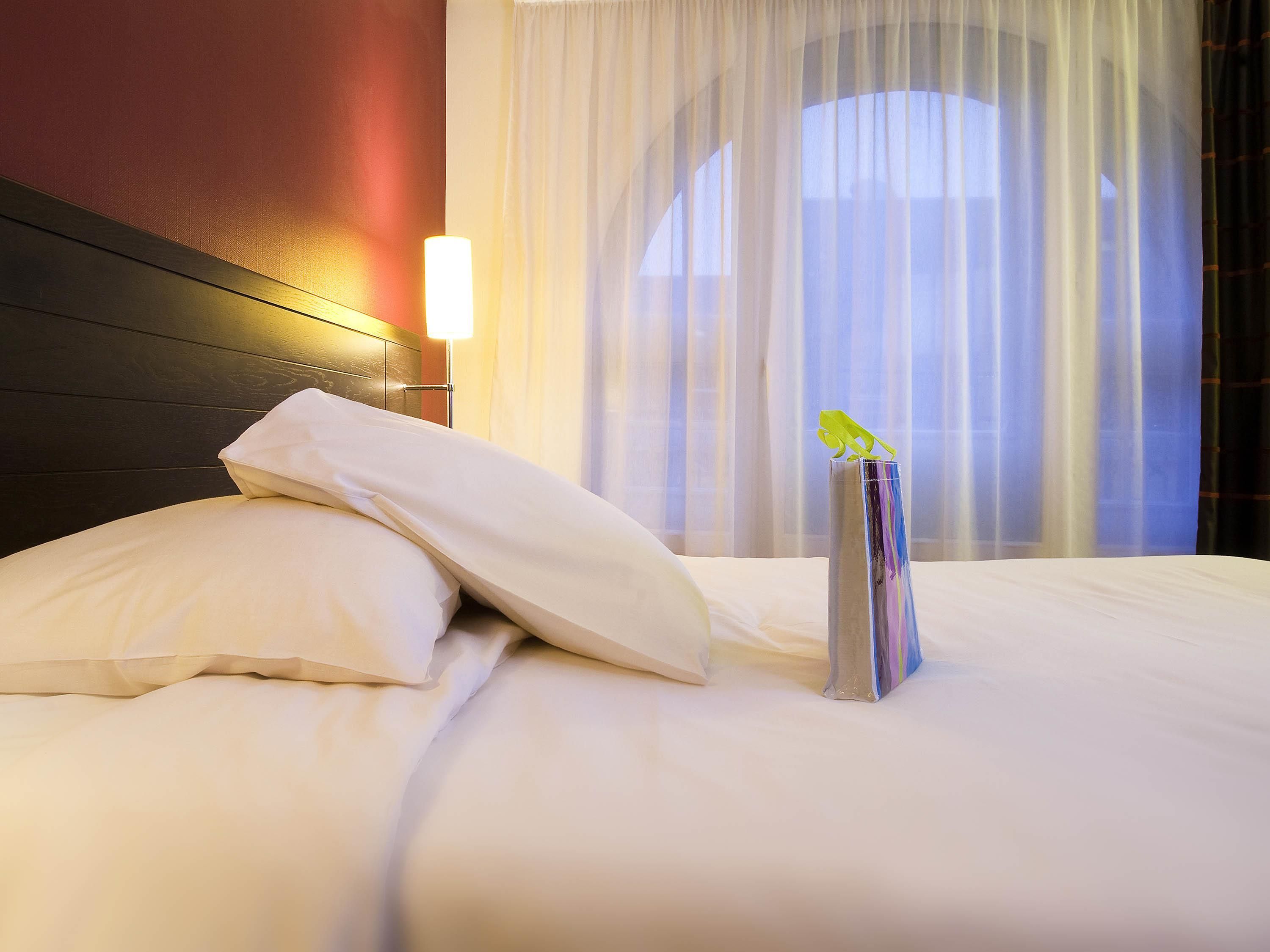 A hotel room with a neatly made bed, a bedside lamp, and a window with sheer curtains.
