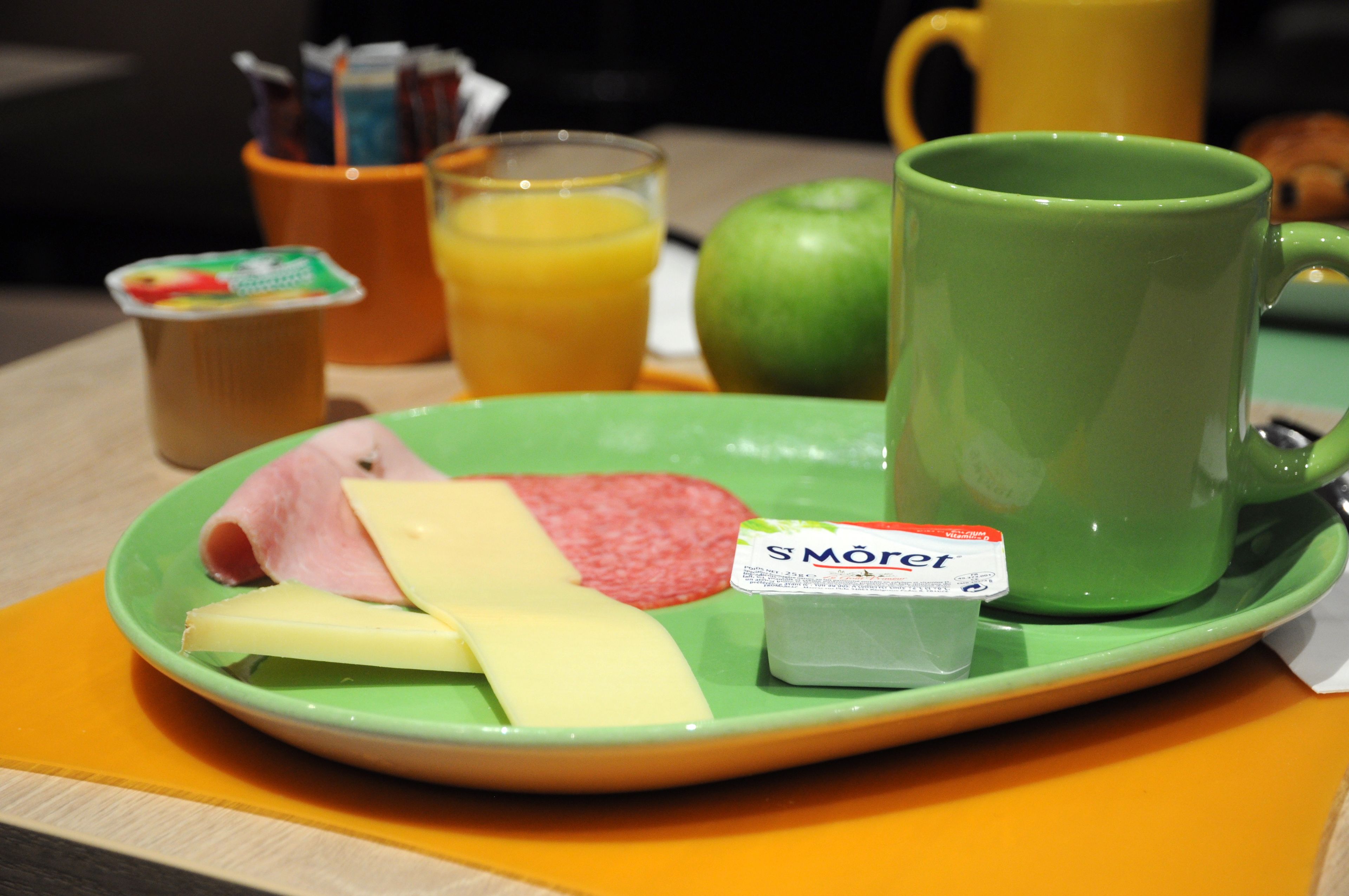 Una bandeja de desayuno con jamón, queso, una manzana verde, un vaso de jugo de naranja, una taza verde y un recipiente con yogur.