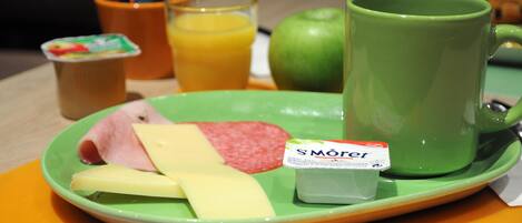 Una bandeja de desayuno con jamón, queso, una manzana verde, un vaso de jugo de naranja, una taza verde y un recipiente con yogur.