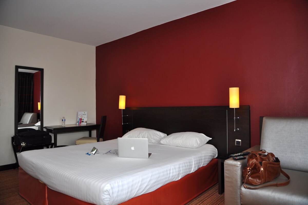 Habitación de hotel con cama, escritorio, silla, ordenador portátil, bolso de mano y mesita de noche con lámpara.