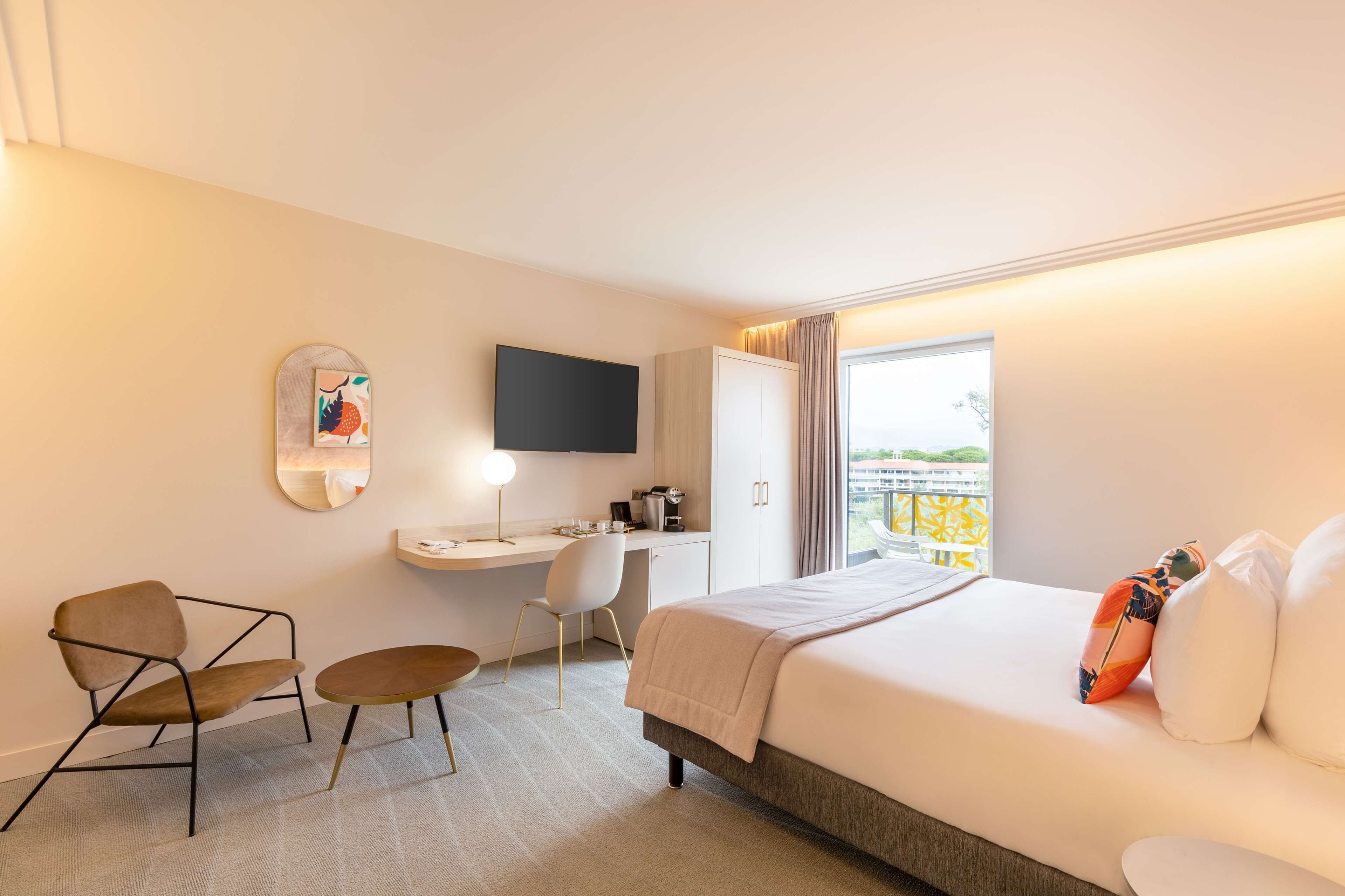 Une chambre d’hôtel équipée d’un lit, d’un bureau, d’une chaise, d’une petite table, d’un téléviseur et d’une fenêtre donnant sur un paysage agréable.